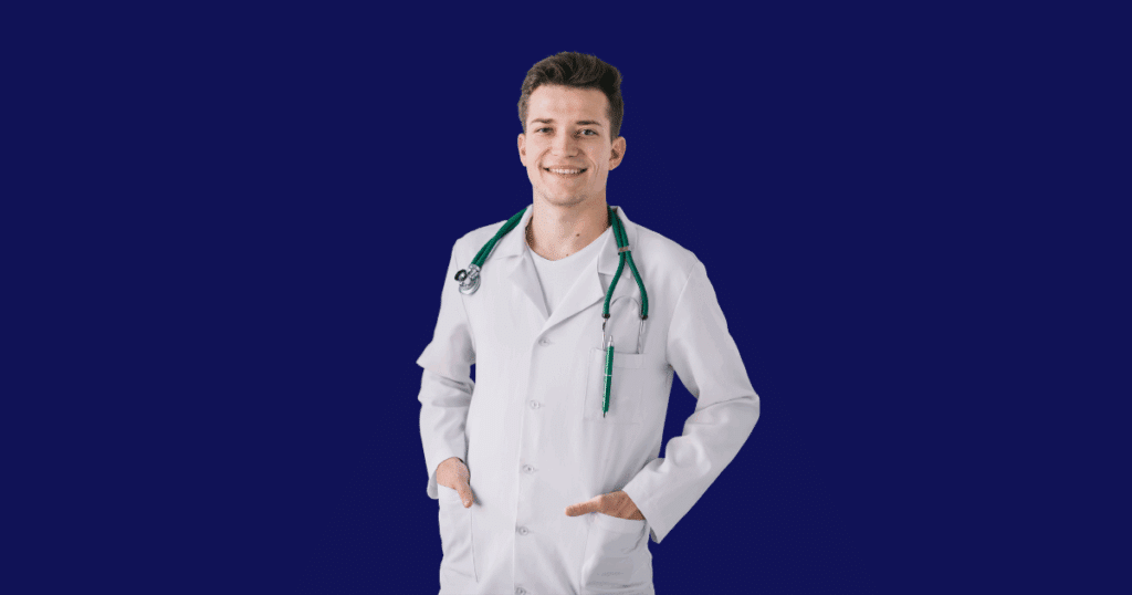 Médico jovem sorridente com jaleco branco, estetoscópio verde no pescoço e mãos nos bolsos, posando em frente a um fundo azul escuro sólido.