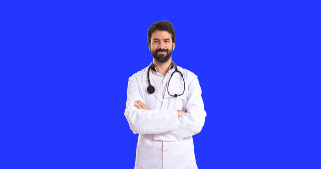 Médico sorridente com barba, de braços cruzados, vestindo jaleco branco e estetoscópio no pescoço, posando em frente a um fundo azul vibrante.