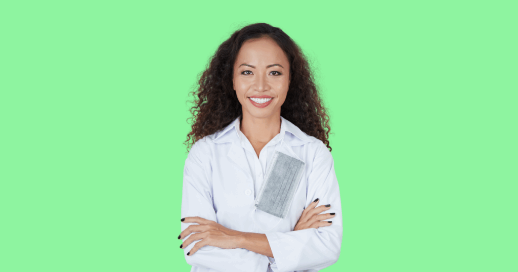 Médica sorridente de braços cruzados, vestindo jaleco branco com uma máscara cinza pendurada no bolso, em frente a um fundo verde-claro.