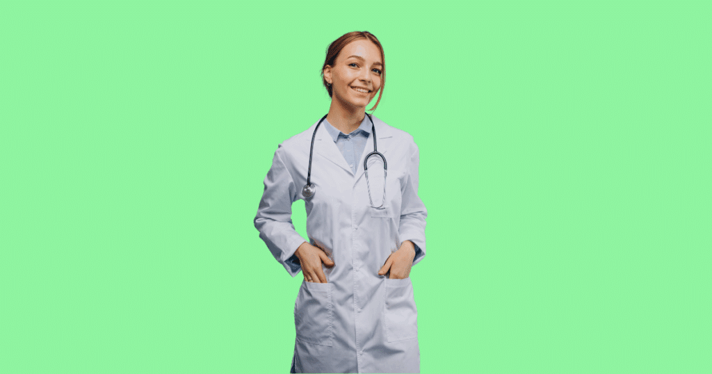 Médica jovem sorrindo, com as mãos nos bolsos do jaleco branco e estetoscópio no pescoço, em fundo verde claro.
