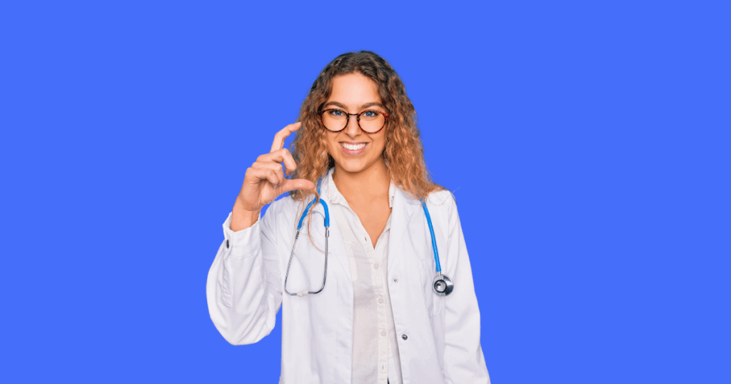 Uma médica loira sorridente, de óculos e estetoscópio, faz um gesto com os dedos, indicando algo pequeno.