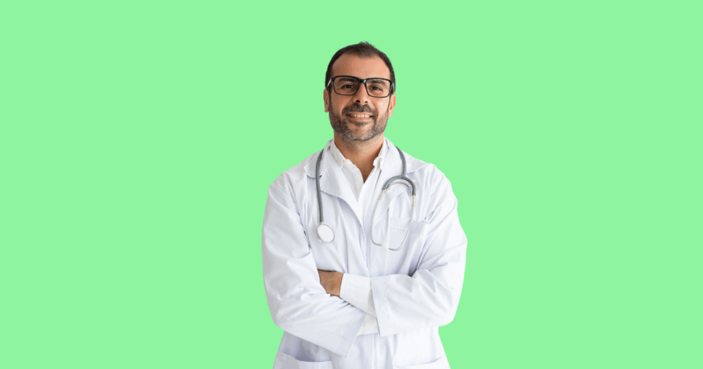 Um médico de barba e óculos, usando jaleco branco e estetoscópio, sorri com os braços cruzados.