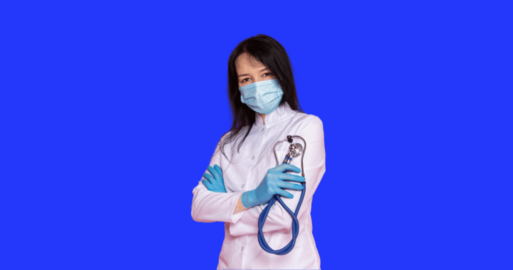 Uma médica de cabelos escuros, usando máscara, luvas azuis e jaleco branco, segura um estetoscópio com os braços cruzados.