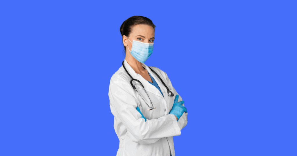 Uma médica com jaleco branco, estetoscópio e luvas azuis, usando uma máscara médica azul, olha para a frente com os braços cruzados.