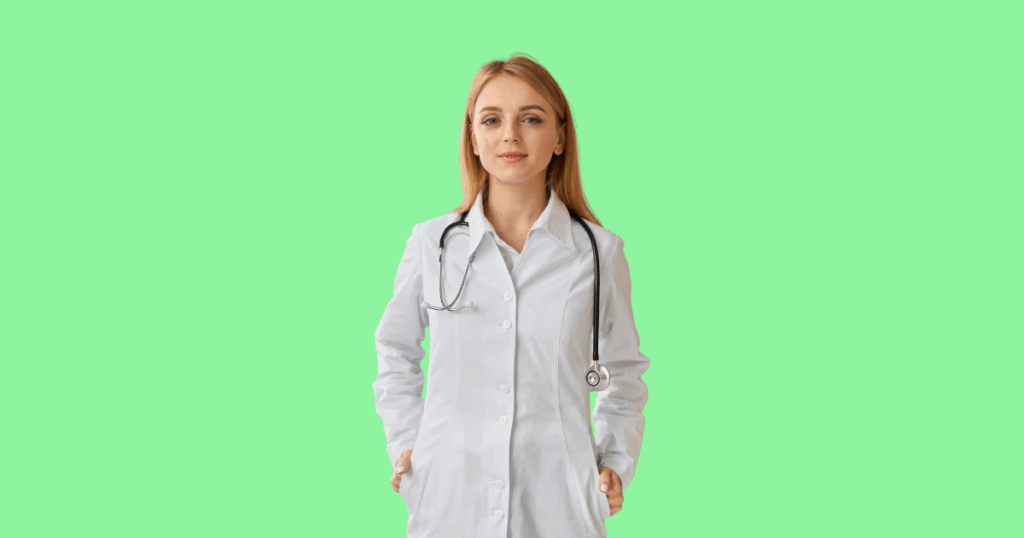 Uma médica jovem e loira, com um jaleco branco e estetoscópio, está com as mãos nos bolsos e olhando para a câmera.
