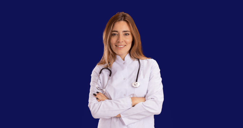 Médica sorridente vestindo jaleco branco e estetoscópio cruza os braços enquanto olha para a câmera. Fundo azul escuro sólido. Imagem ilustrativa para conteúdo sobre tipos de tributos no Brasil.